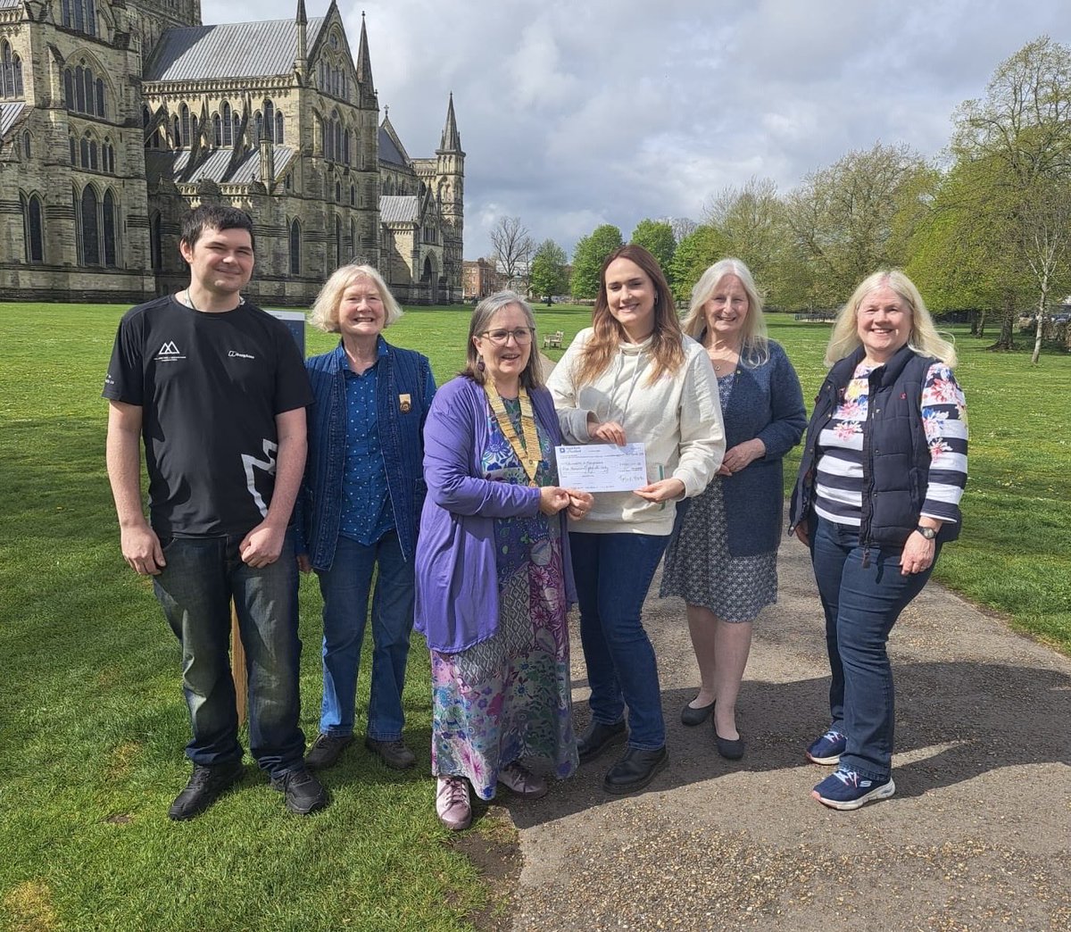 Cindy D-G, Chairman 2025-26 proudly presenting a cheque for £1000.00 to Kimberley from Millimetres 2 Mountains, charity funds raised by Salisbury Tangent (389) with Liam (recipient of support), Jocelyn P, Chairman 2026-27 &amp; members Jacky W &amp; Caren C by Salisbury Cathedral