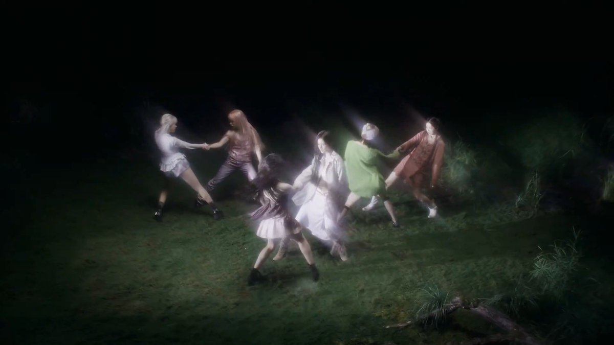 Six female performers holding hands and dancing in a circle in a dark, misty outdoor setting with grass, lit from above.