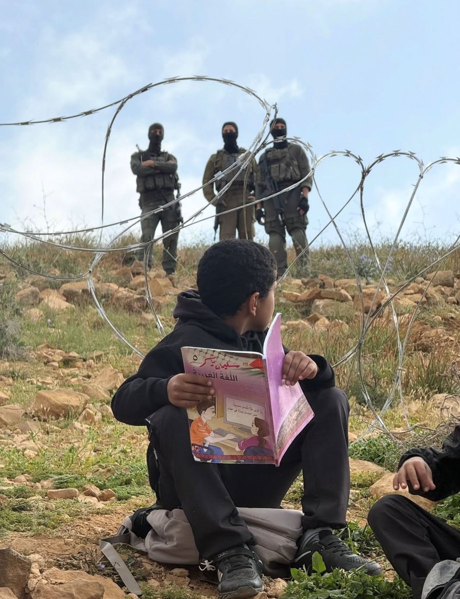 Un niño palestino lee su libro educativo infantil ante la vigilancia de soldados sionistas, detrás de una valla de púas, puesta por los colonos para que no pueda ir a la escuela en la aldea de Umm al-Khair, en Cisjordania.

Una foto que define el apartheid sionista y que bien
