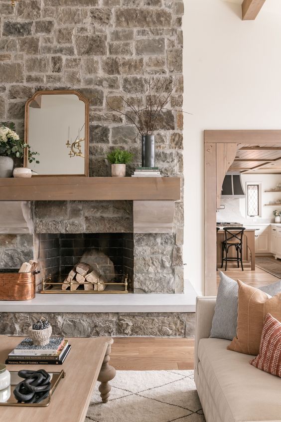 Textured stone fireplace with a wood mantel creates a warm, modern rustic living space. A strong example of natural stone masonry, neutral interior design, and functional open-concept layout.
 arnoldmasonryandlandscape.com