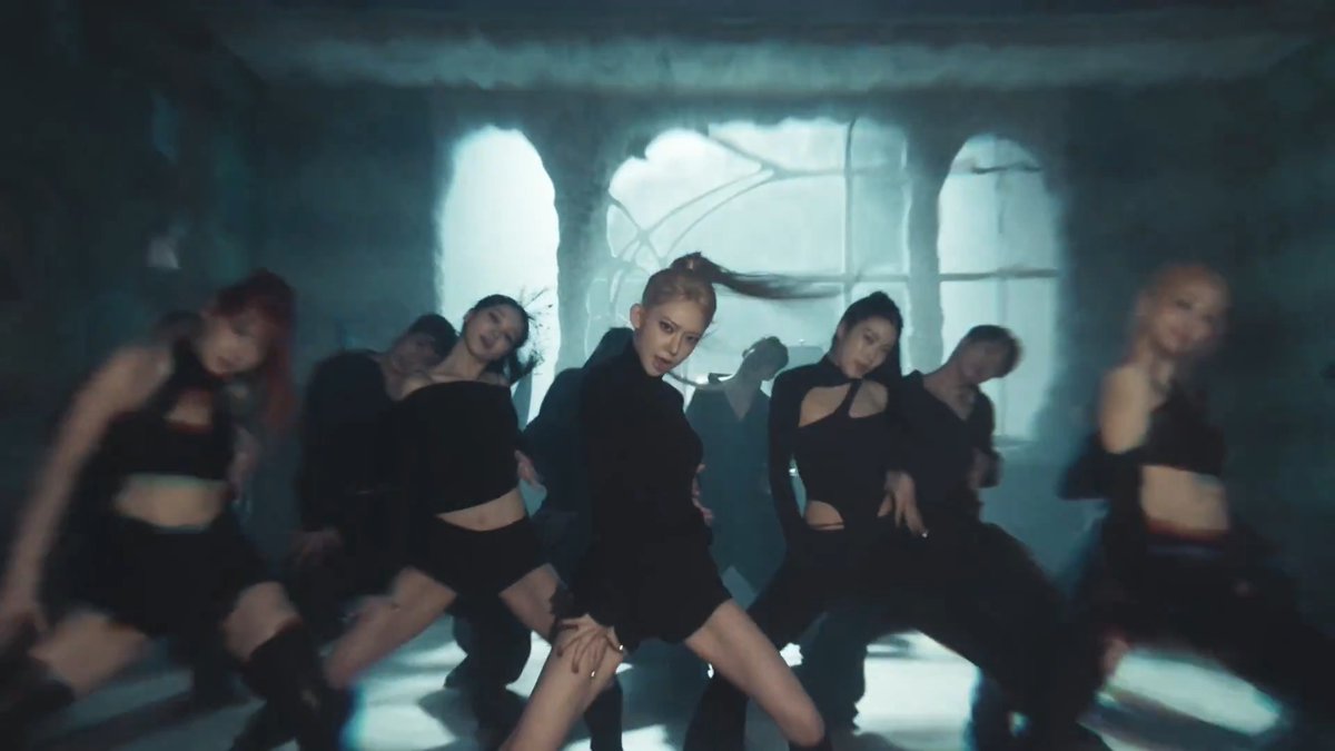 A group of women in black outfits perform a choreographed dance in a dimly lit, atmospheric room with a large arched window.