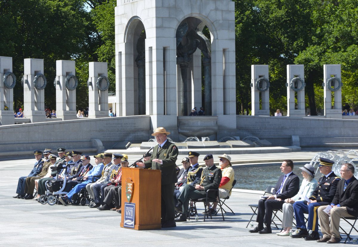 A park ranger in uniform and hat speaks from a podium with a line of dignitaries seated behind him in an outdoor military memorial plaza on a sunny day.