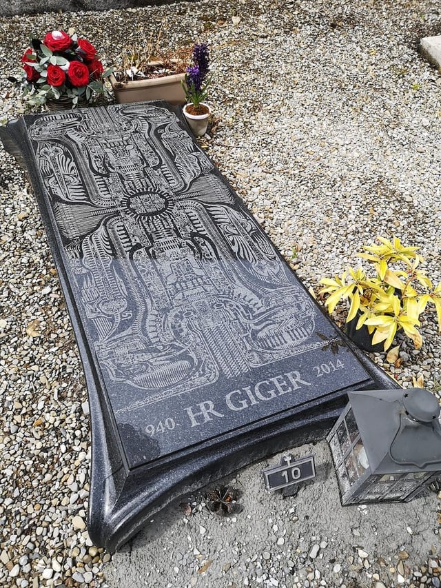 The grave of legendary Swiss surrealist artist H.R. Giger.
Famous for his biomechanical style and designing the Xenomorph,  lies in Cimetière de Gruyères, a short walk from the H.R. Giger Museum and Giger Bar in the medieval town of Gruyères, Switzerland.