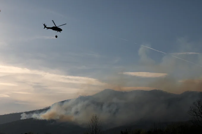 Helicopter carrying a water bucket flies over smoky mountains to combat a wildfire at dusk.