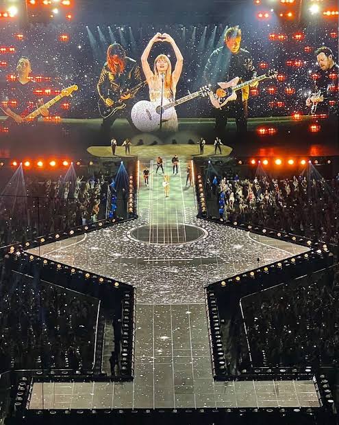 uma loira com um telão gigante coberta de gliter tocando instrumentos com sua banda e se divertindo no palco eu acho que tô tendo alucinações