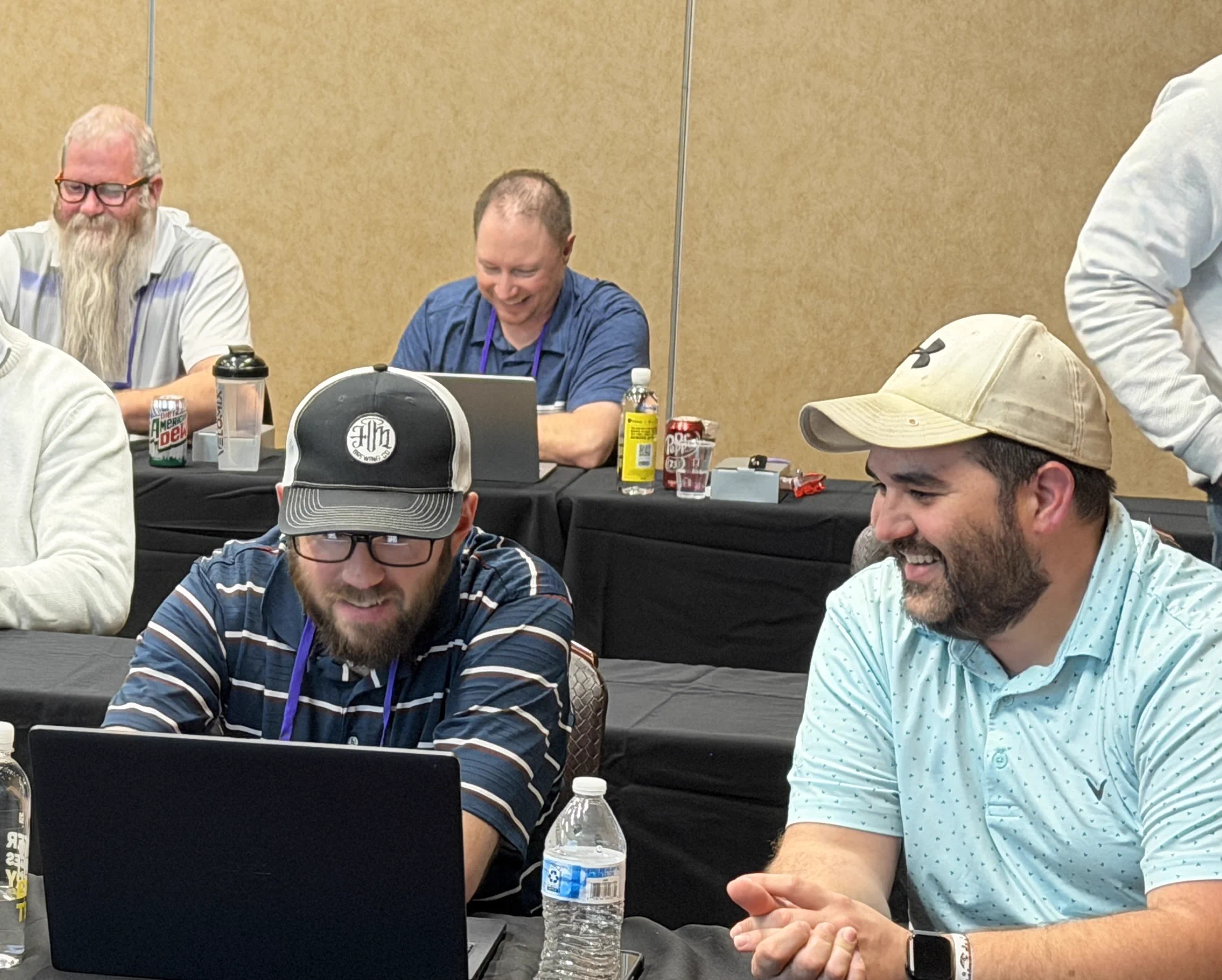 A group of people engage in a collaborative workshop, with some smiling and working on laptops. The atmosphere is relaxed and positive.