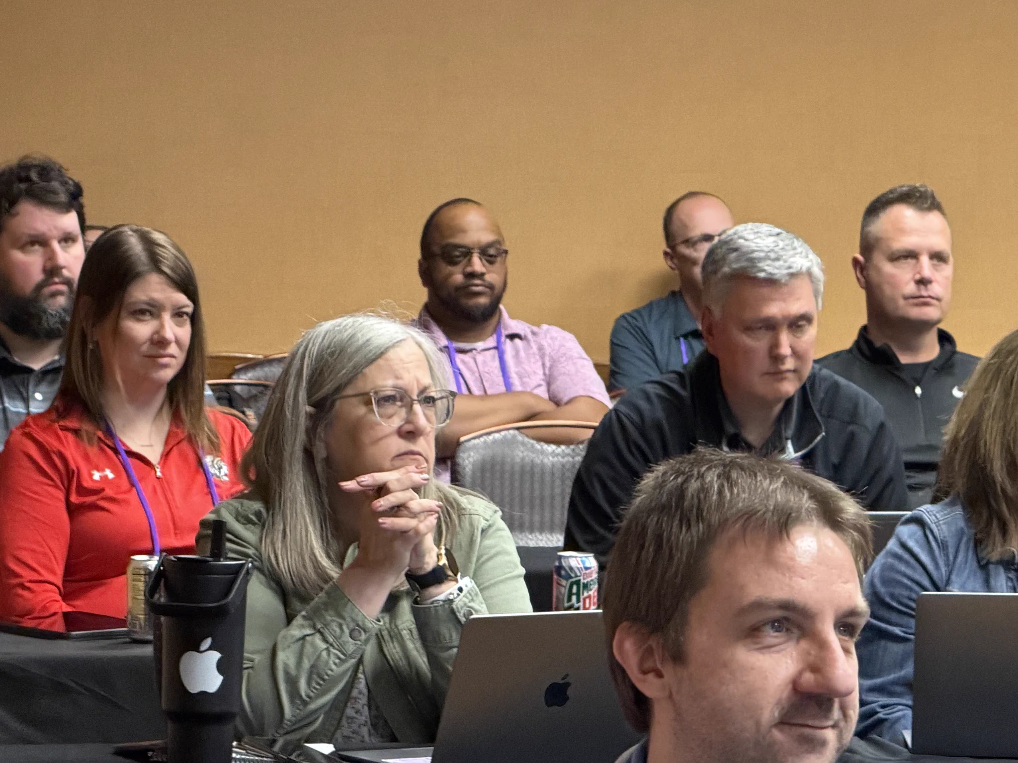 A group of individuals attentively listen during a conference session. The setting appears professional and focused, with laptops open and note-taking in progress.