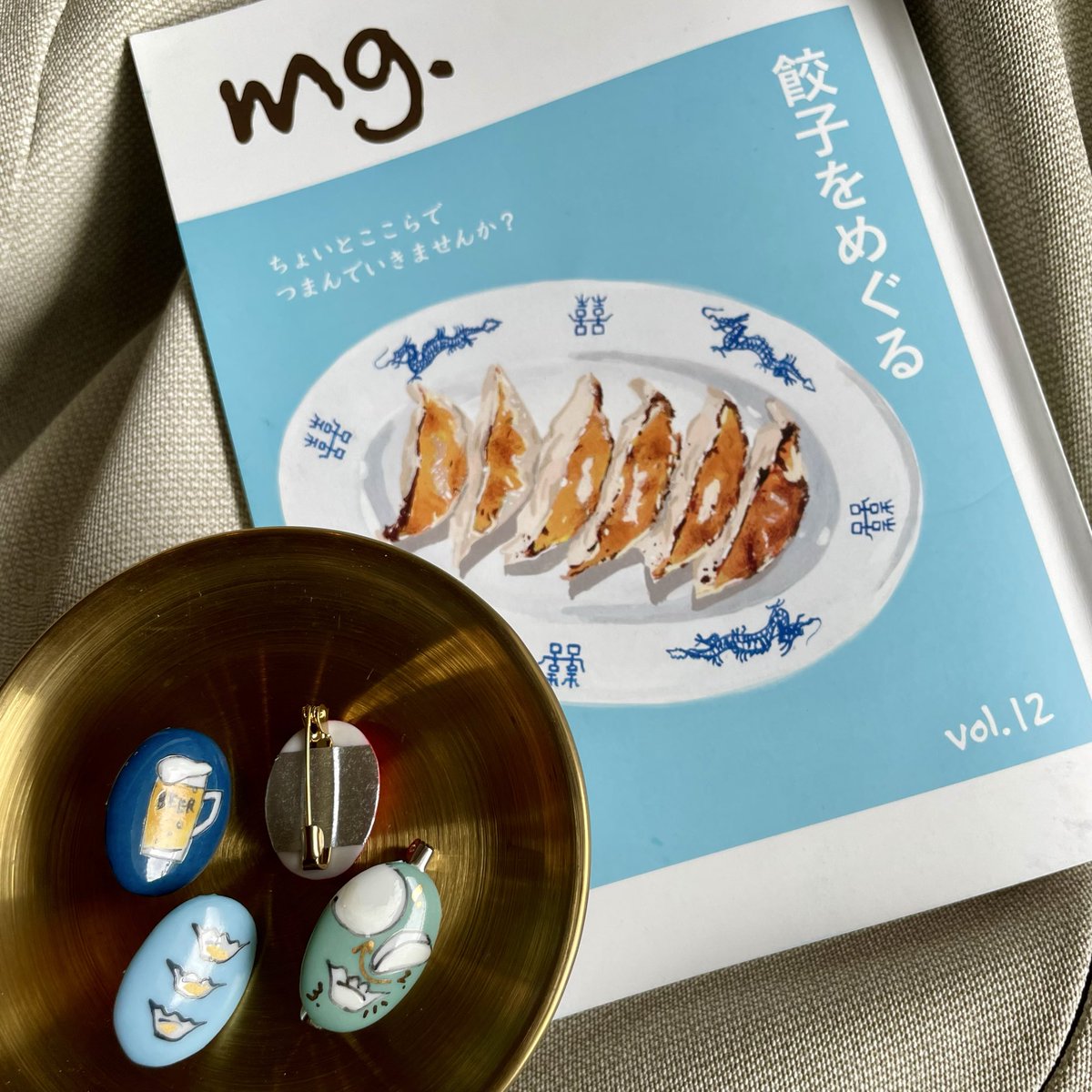 やはり表紙合わせの水色餃子🥟と🍺を耳に！残りはブローチに。これ、実は右上パーツのように裏でマスキングテープで付けています！接着剤だと乾燥時間が足りなくて。白磁がとても軽いので、今日のように1日だけの使用のときはこれでも保つんです✌️【こ83-84】にて、ぜひ実物ご覧ください🩵