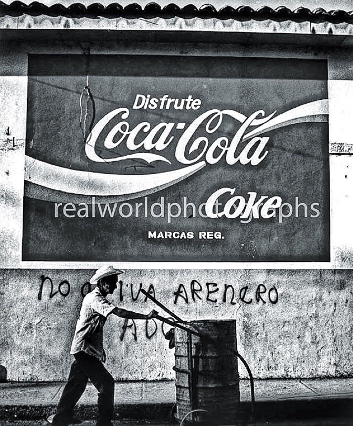 RealWorldImage's tweet image. A garbage collector walks under an advertisement for Coca-Cola in San Salvador, El Salvador, Central America. 1993. Gary Moore photo. Real World Photographs. #streetphotography #photojournalism #CocaCola #Grok  #Nikon #ElSalvador #garymoorephotography #realworldphotographs