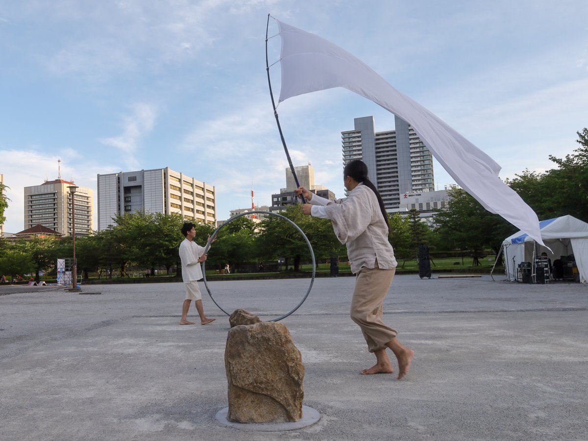 masato009's tweet photo. うきも-project-『うきふね』
2026年5月4日(月)17:30〜18:00
駿府城公園富士見ひろば

大熊隆太郎『末待奉祭2026』に大いに満足し、すぐ隣の富士見ひろばへ。次は「うきも-project-」。まったく知らない。見ることになったのは、時間と場所が『王女メデイア』までのつなぎとして良かったからだ。

ところがこれが実に良かった。まさに不意打ちのような素晴らしさだった。

今年はSHIZUOKAせかい演劇祭もストレンジシードも明らかにスケールダウンしていた。ストレンジシードについて言えば、以前と比べれば出演アーティストが半分くらいに減ってしまい、２泊３日でも全部は見切れない、あのオモチャ箱をひっくり返したような楽しさと熱気が失われていた。
しかしたった３つしか見なかった演目、コレクティフ プロトコル『One Shot』、大熊隆太郎『末待奉祭2026』、うきも-project-『うきふね』が、どれも実に見事なパフォーマンスだった。SHIZUOKAせかい演劇祭では、バロ・デヴェル『Qui som?─わたしたちは誰？』には不満を覚えたが見る価値はあった。SPAC『王女メデイア』は言わずもがな。演目で言えば、２日間でたった５つしか見られなかったが、「平均点」という視点からすれば、これまででも屈指の年だった。確かに数は減ったし、その意味ではスケールダウンなのだが、実は非常に「少数精鋭」で、今回見られなかった演目も充実したものだったのでは？と思えてきた。

うきも-project-はシルホイールという大きな輪と巨大な白旗を使ったパフォーマンス。その動きは時にはサーカス的、時には現代アートのようであり、時には宗教儀式のようでもある。その３つの間を行ったり来たり。最後は舞台に置かれた「石」が意味を持つあたり、先ほどの『末待奉祭2026』とは違う形で日本の神道に通じるものを感じた。ニューエイジっぽい音楽もそこにピッタリはまっている。

このプロジェクトは香川から来ているそうで、海辺などいろいろな場所で上演しているらしい。香川までは見に行けないので、なかなか見る機会はないと思うが、近場で見られるときは必ず見ようと思う。パフォーマンスだけでなく、風景が重要な意味を持つので、仮に内容が同じでも、場所が変われば別物になることだろう。人と風景が一体となって生まれる稀有なパフォーマンス。大熊隆太郎に続いて、本当に良いものを見せてもらった。最初に述べたように、今年はアーティストの数こそ激減したが、その分 少数精鋭だったとしか思えない。

ところでこれらの写真、前の大熊隆太郎に続き、うっかりブラックミストフィルターを装着したまま撮ったら、撮影対象が白基調かつ薄曇りの光でフィルターとの相性が最悪。家で見たら、全部「眠い」写真になっていて愕然とした。
仕方なくレタッチでホワイトバランスまで変えて、ようやく少しはマシなものにしたが、アーティストから「できるだけ肉眼で見て欲しい」と言われていたこともあり（その意図はよく分かる）、何となく思いきりの悪い写真ばかり。もう少し写真の腕を磨きたいなあ…(´-ω-`)

#ストレンジシード静岡2026
#うきもproject
#うきふね