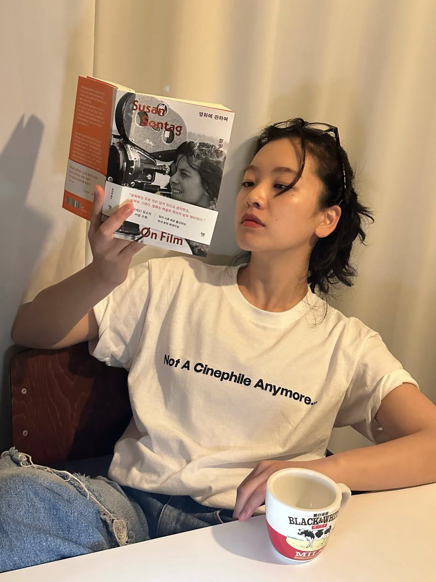 deanfluencer's tweet photo. actress lee jooyoung stuns in a “not a cinephile anymore...” shirt, reading susan sontag on film book and drinking from a black &amp; white tea cup (iconic hong kong milk brand) i love her so much! https://t.co/l5KPtS0aZw