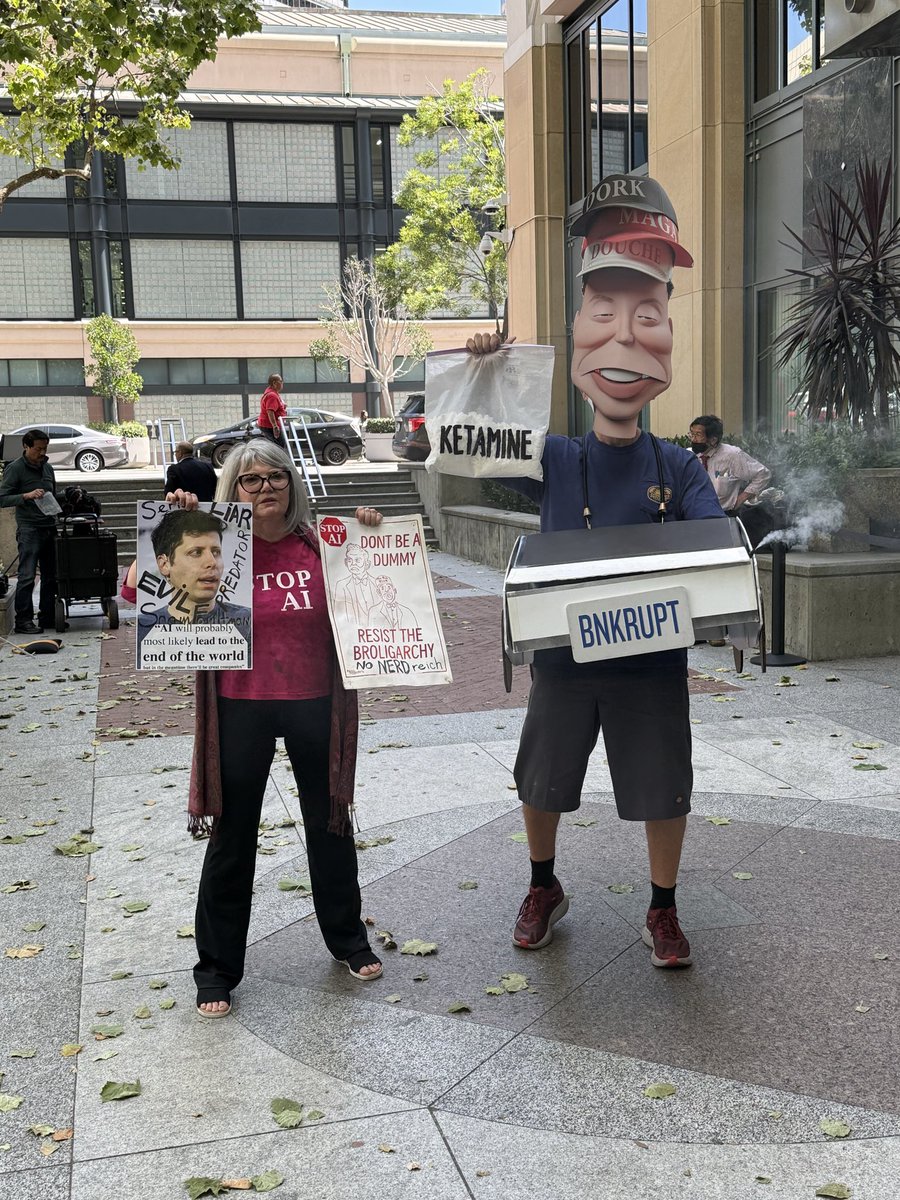 michelletomkim's tweet photo. In front of the Oakland federal courthouse, a protestor wears an Elon bobblehead, holding a bag of ketamine and a cybertruck prop, with an inflatable tube man imitating Musk in a MAGA cap. Another protestor holds a photo of Sam Altman saying “serial liar.” https://t.co/z4Z3OVwgoy