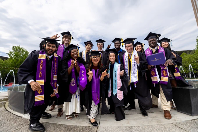 A group of graduates wearing caps and gowns celebrates in front of a fountain. They are holding diplomas and wearing stoles with Greek letters. Trees and a cloudy sky are in the background.
