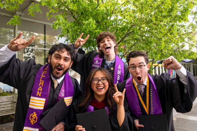 Four graduates in gowns and purple stoles celebrate outside, smiling and holding their caps. Trees and a building are in the background.