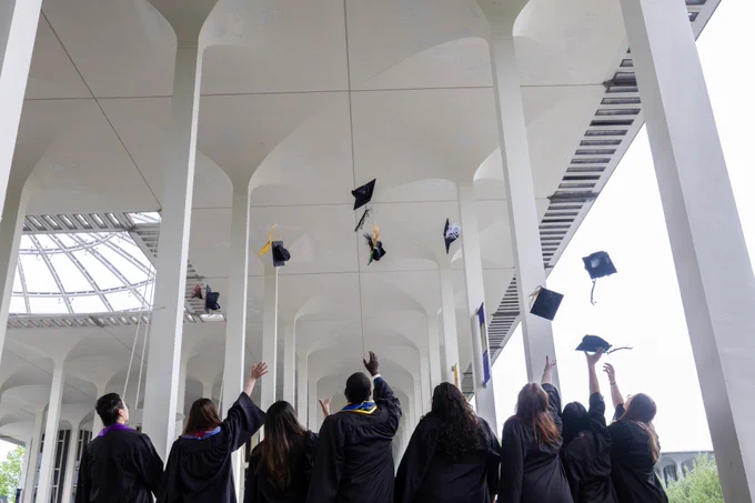 A group of graduates in black gowns celebrate beneath an architectural structure at the University at Albany by throwing their caps into the air.