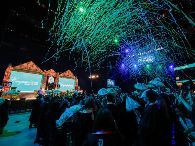 Graduates celebrate at a commencement ceremony in a large indoor venue, the Caesars Superdome. The stage displays "Congratulations Class of 2026," and colorful streamers fall from above, creating a festive atmosphere.