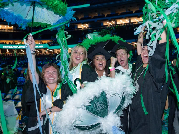 A group of Tulane University graduates in robes celebrate enthusiastically, holding decorative umbrellas in a vibrant stadium setting. The background displays a congratulatory message for the Tulane University Class of 2026.