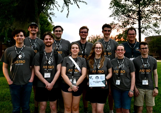 Group of eleven Michigan Tech students wearing matching Astro Huskies shirts posing outdoors with one holding a certificate.
