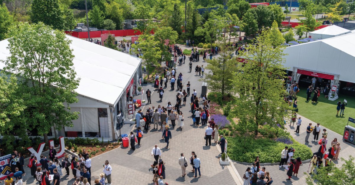 Aerial view of a busy outdoor event with people walking and mingling on a large paved pathway lined with trees and greenery. There are two large white tents and several smaller red structures visible. The scene appears vibrant and lively.