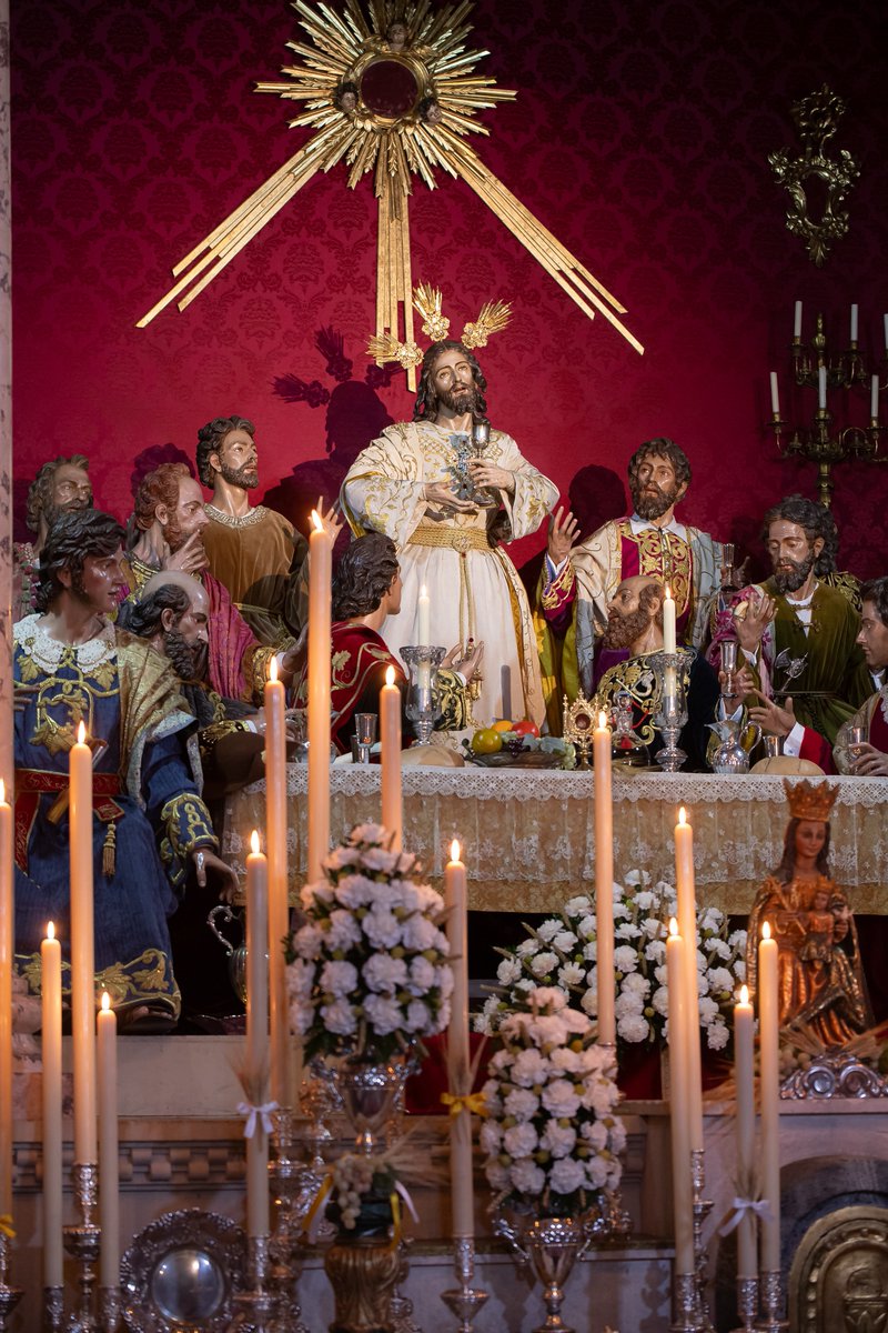 lacenademalaga's tweet photo. #CorpusSagradaCena26 - El Señor de la Sagrada Cena se presenta junto al colegio apostólico en su capilla, especialmente remozada para la celebración del triduo eucarístico, que tendrá lugar los días 4, 5 y 6 de junio de 2026, a las 19:00 horas, en la iglesia parroquial de Santo Domingo de Guzmán.

La hornacina que alberga el conjunto escultórico ha sido revestida de damasco. Sobre el arco de la capilla se dispone un amplio cortinaje que actúa como marco escenográfico y sobre el que se abre visualmente toda la composición. Se completa con dos cornucopias, dos candelabros y un gran sol radiante cuyos rayos descienden sobre la imagen del Señor, reforzando la centralidad de Cristo como fuente de luz y salvación.

Para esta ocasión, el Señor se presenta sosteniendo el cáliz, evocando el momento en el que el sacerdote lo eleva tras la consagración del vino. Este gesto subraya el profundo misterio de la fe cristiana. Debido a la visita del Santo Padre a España, el Señor luce una estética dominada por los tonos blanco y dorado, colores estrechamente vinculados al pontificado y al propio Vaticano. Asimismo, el altar incorpora guiños a este acontecimiento excepcional, como los lazos blancos y amarillos que sujetan las espigas dispuestas en los cirios.

Cada apóstol aparece acompañado de sus atributos iconográficos. Así, por ejemplo, Santiago el Mayor porta el sombrero de peregrino y con la concha jacobea, símbolo de su vinculación con las peregrinaciones compostelanas; San Pedro aparece representado con las llaves, atributo que recuerda su condición de primer pastor de la Iglesia y depositario de las llaves del Reino de los Cielos; mientras que San Judas Tadeo sostiene el hacha, instrumento asociado a su martirio según la tradición cristiana. En la mesa sobre la que se dispone el misterio se sitúa un relicario que custodia una reliquia vinculada al cenáculo del Señor. El conjunto se enriquece con diversos motivos de marcado simbolismo eucarístico con el que propio apostolado interactúa.

El exorno floral está compuesto por helecho, claveles, uvas y espigas.

📸@j16photos

#CofradíasMLG