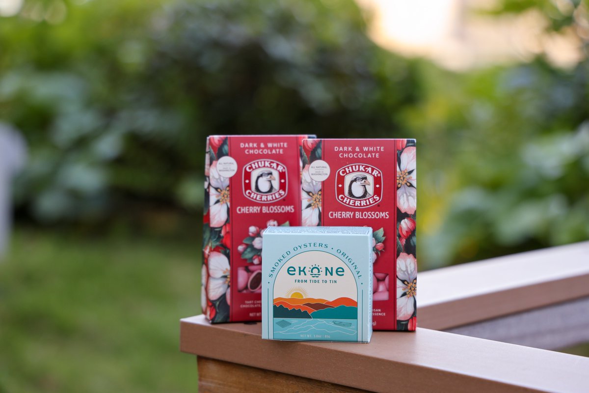 Chocolate products from Chukar Cherries and a package of Original Ekone Smoked Oysters from Taylor Shellfish Farms are displayed outdoors against a background of lush greenery.