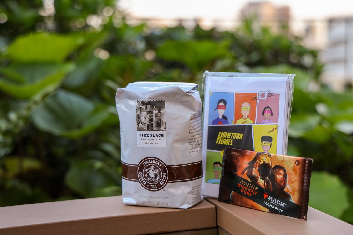 A package of "Pike Place" roast coffee from Starbucks, a set of colorful "Hometown Legends" illustrated cards, and a "Magic: The Gathering" card pack are displayed on a table with a leafy background.