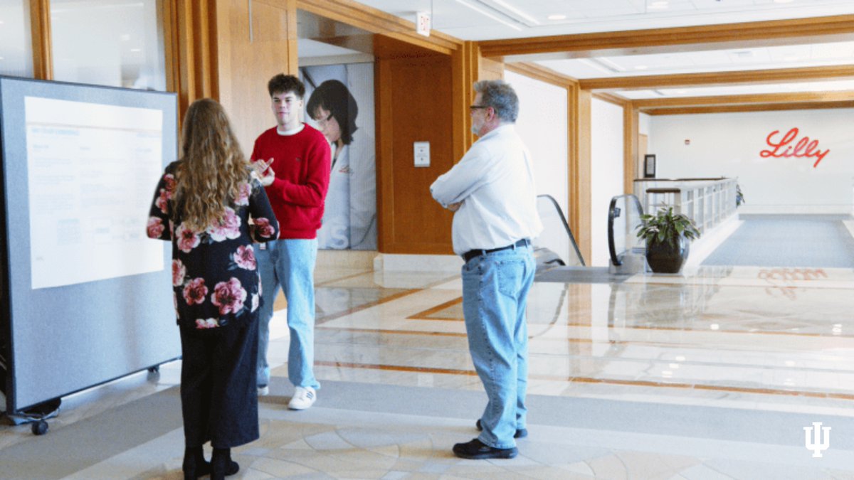 Three individuals are engaged in a discussion in a well-lit hallway near an informational display. The space features a visible Lilly logo on the wall, indicating the location. The setting has a professional atmosphere with polished floors and wooden accents.