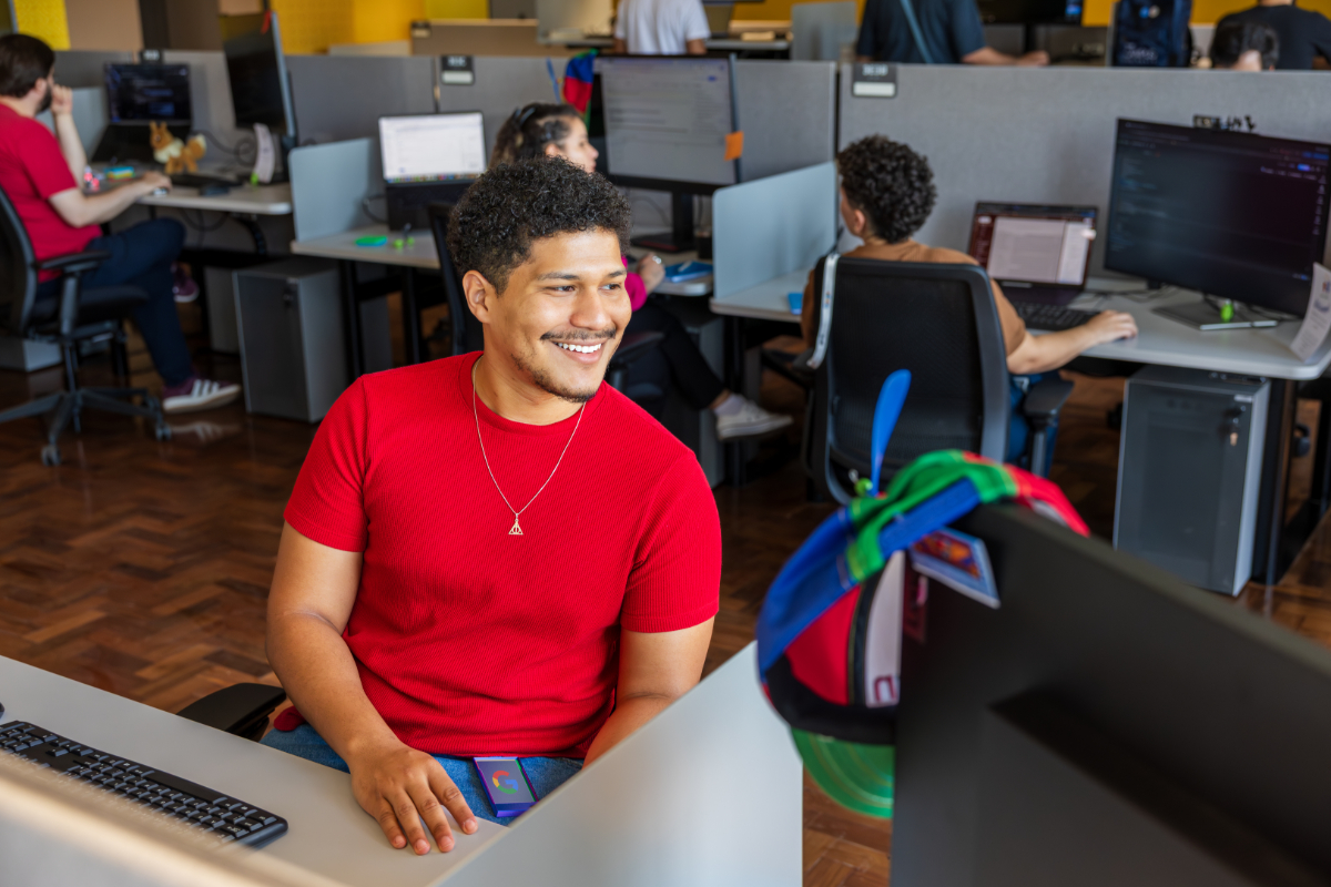A man in a red T-shirt sits at his desk smiling, surrounded by other coworkers working at computers in an open-plan office space.