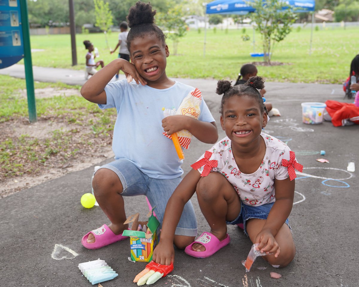 Two children playing drawing with chalk on the pavement smiling at the camera.