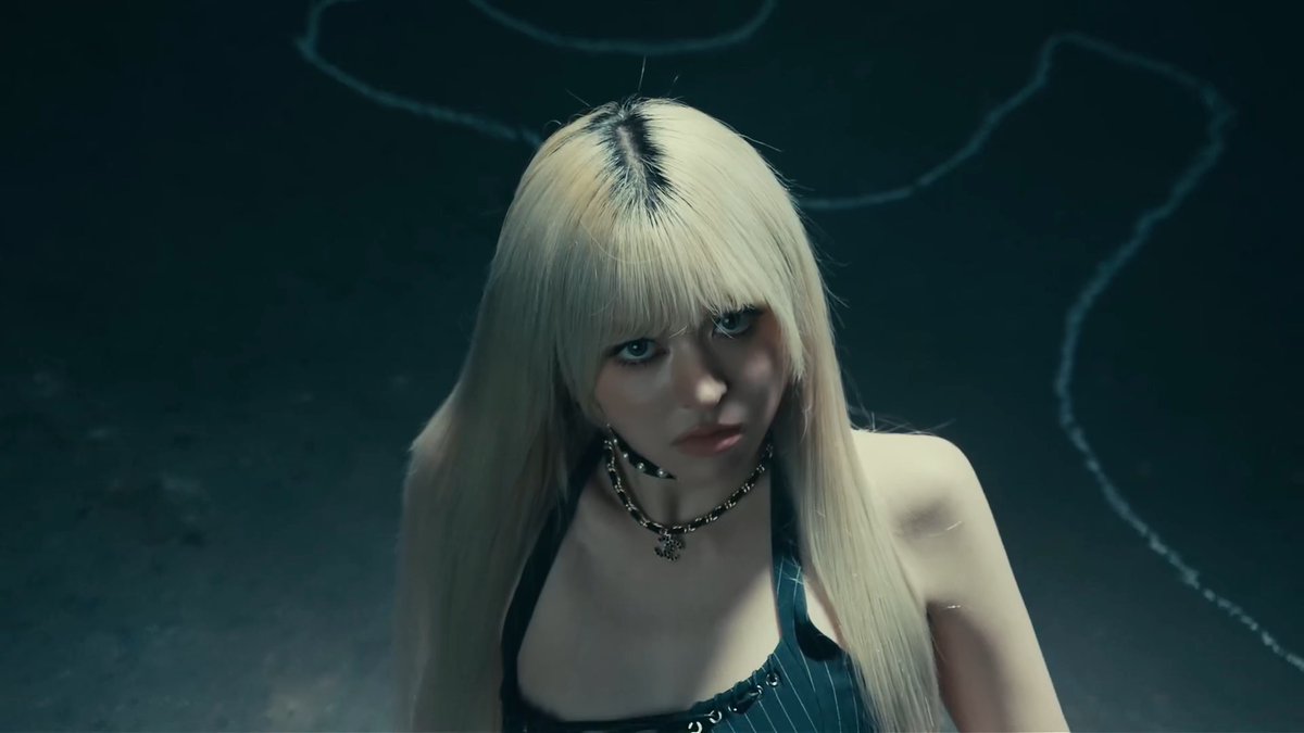 A performer with platinum blonde hair and dark roots wears a black choker, spiked collar, and striped top against a dark background.