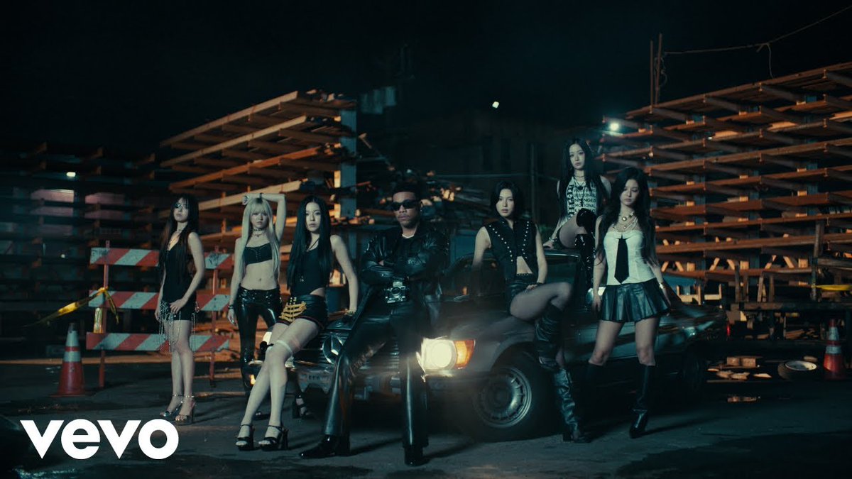A still from a Vevo music video showing seven women in edgy black outfits posing in front of a car in a construction yard setting.
