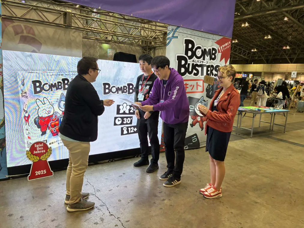 EngamesToyama's tweet photo. 「世界のボードゲームを広める会 ゆうもあ」様が主催する「日本ボードゲーム大賞2025」の投票部門でボムバスターズが最多得票数となる大賞を授賞しました。
授賞を記念して、主催者様よりゲームマーケット会場にて代表・杉木とゲームデザインを担当された林尚志さん（OKAZU brand）に賞状をいただきました。
販売元であるCocktail GamesのJulia KLOKOVAさんにも同席していただきました。