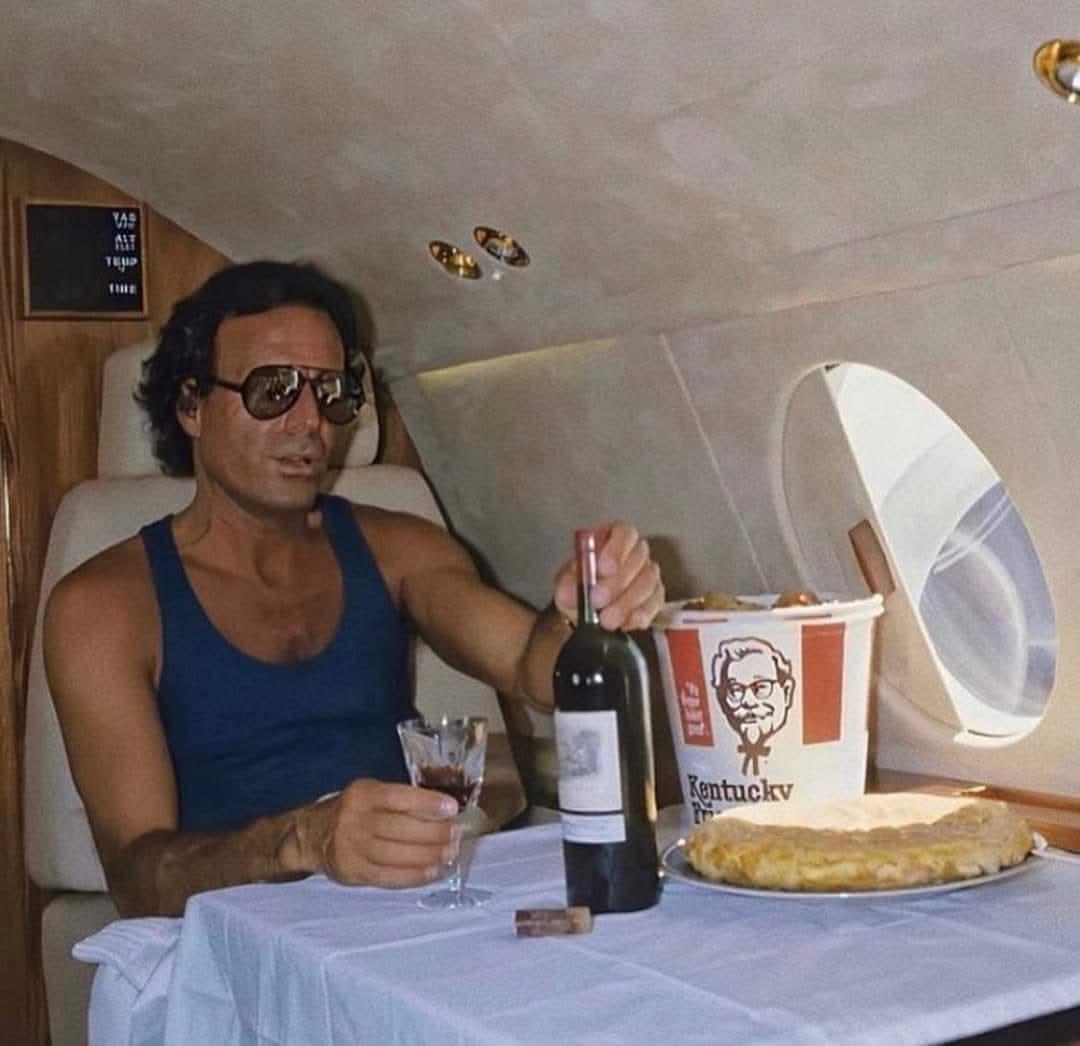 HistorylandHQ's tweet photo. Julio Iglesias enjoying a KFC family bucket and tortilla de patatas with a bottle of Château Lafite-Rothschild on his jet. Photograph by Jean-Claude Deutsch (1986) https://t.co/gze7IAIrNX