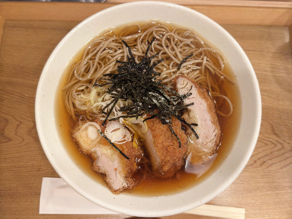 akabanemagazine's tweet photo. 赤羽ララガーデン「中村麺兵衛」で食べた『とんかつ出汁そば』。

お蕎麦にとんかつ！？というなかなか攻めた組み合わせにワクワクしながら待っていると、運ばれてきたのは存在感たっぷりの一杯。

お箸で持ち上げると、ずしっと伝わる重量感！そのままお出汁にたっぷり浸して、衣にじゅわっと吸わせてからがぶり。

おっ、お肉柔らか〜！これはいいぞ！

とんかつと蕎麦って別々で食べるイメージが強かったけど、出汁をまとったカツが妙にしっくりくるんです。

また、天かすではなく、とんかつの衣がお出汁と絡む感じがなんとも新鮮。