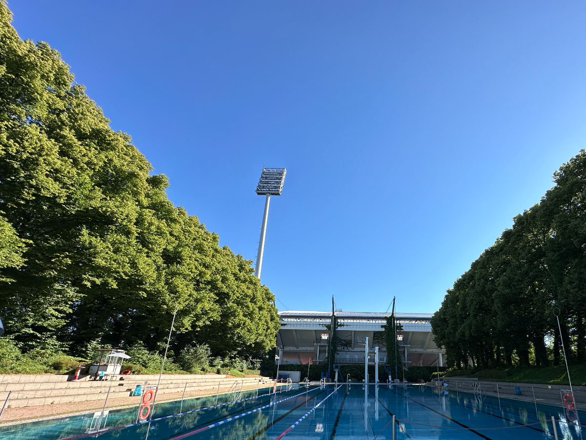 Leeres Freibadbecken vor dem Stadion, flankiert von hohen Bäumen unter klarem, blauem Himmel.
