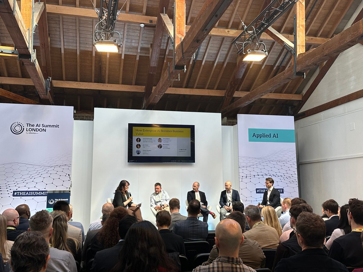 A panel discussion at The AI Summit London. Five speakers, including one woman holding a microphone, sit on white chairs on a stage. A screen displays \"How Enterprise AI Becomes Business Value.\" An audience sits watching. Banners read \"The AI Summit London\" and \"Applied AI,\" with #THEAISUMMIT.