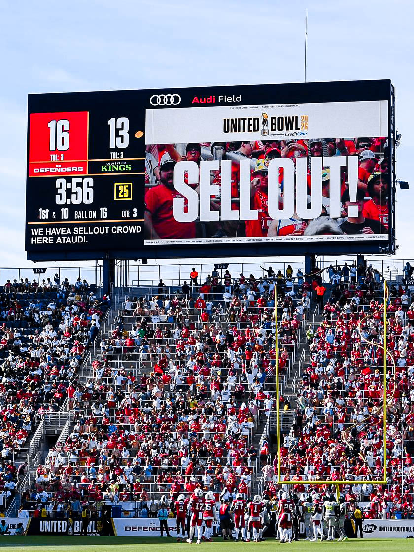 A packed stadium during a football game featuring a massive 'SELL OUT!' sign. Fans in red and white fill the stands, creating an energetic atmosphere.