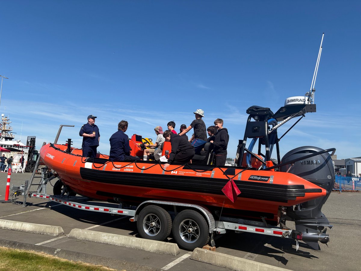 An orange rigid inflatable boat carrying several seated passengers is secured on a double-axle trailer parked near a waterfront. Traffic cones and industrial structures are placed nearby under a clear blue sky.