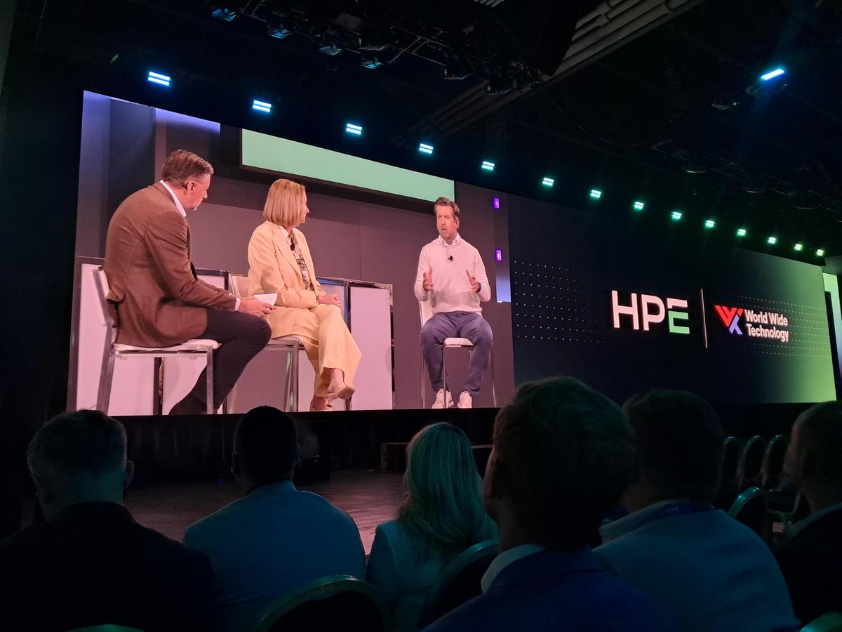 An audience views a stage panel discussion with three speakers. A man in a brown jacket and a woman in a yellow suit sit to the left, while a man in a white sweater gestures as he speaks. Behind them, a large screen displays \u201CHPE\u201D and \u201CWorld Wide Technology.\u201D