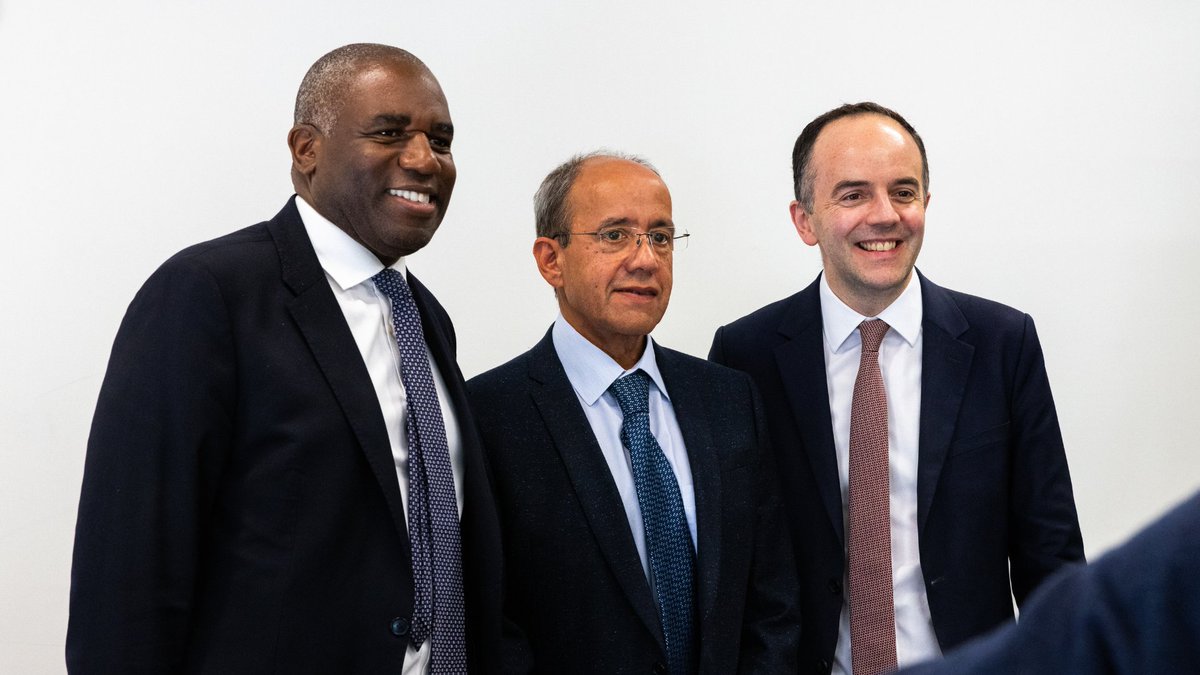 Three individuals in business attire are standing together against a plain background, all smiling.