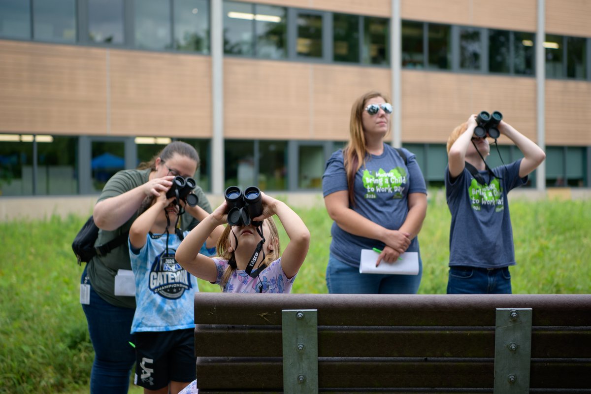 Group of children and adults outdoors using binoculars to observe nature near a modern building.