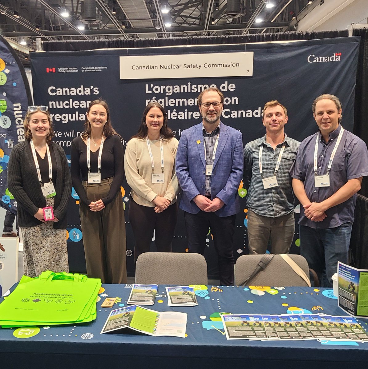 Six CNSC staff members stand behind a conference booth at an indoor conference space, with bilingual signage “Canada’s nuclear regulator / L’organisme de réglementation nucléaire du Canada”, with informational brochures and green tote bags displayed on the table.