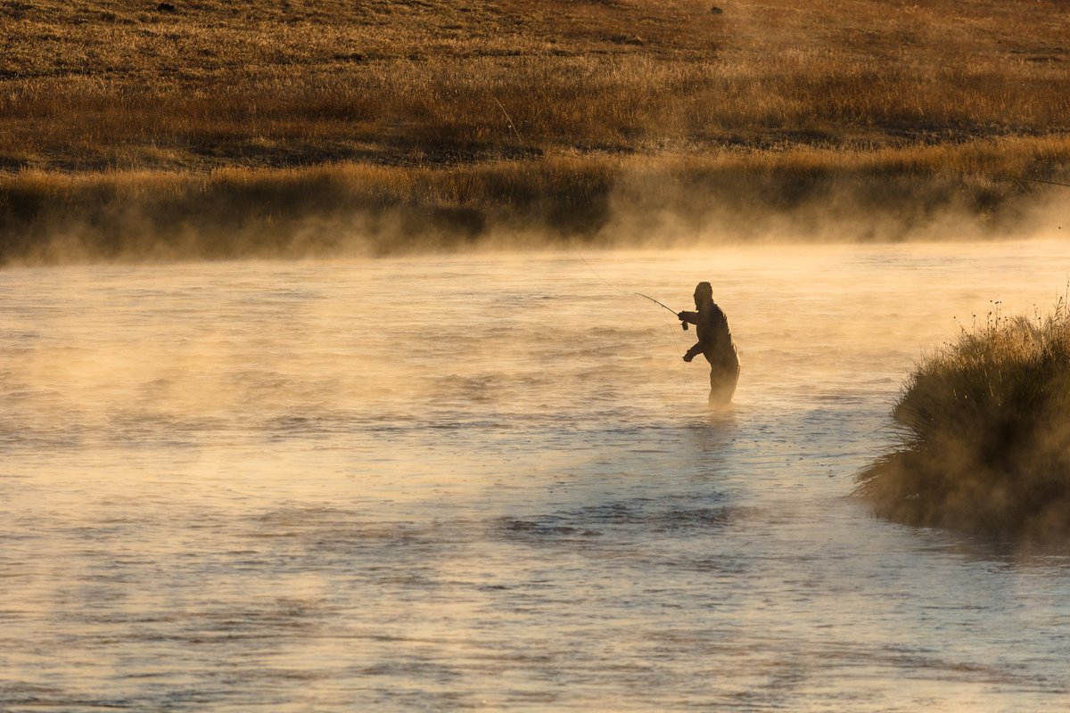 A person standing knee-deep in a river at dawn. The person is casting a fishing line into the water. Steam can be seen rising from the river.