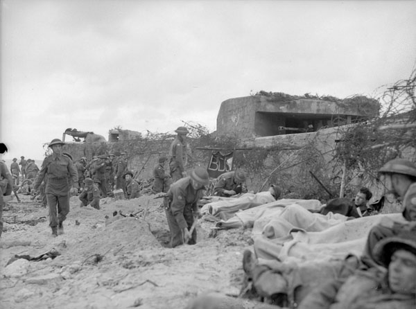 Des militaires en uniforme sur un sol sablonneux, près d’un haut mur de béton surmonté de fil barbelé, en temps de guerre.