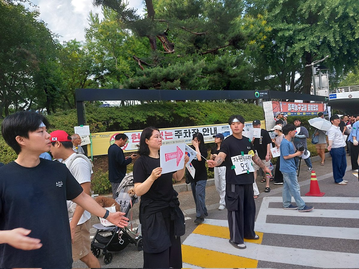 DrTaraO's tweet photo. 2026-6-7. Olympic Park, Seoul. A strange group was controlling the entry to the rally area. They forced people to form a line and go around to enter the area. Why? Who are they? They said they're "spontaneous volunteers," yet they acted like a well-organized group. Someone told them to direct the people to go in the rally area 1 by 1, standing in a line. They claimed its for safety, but I don't see why going into a large open area requires a line. They were forceful about it. One older man (see photo) pushed me to go in a line going in a different direction. I've never seen this. People should be free to come and go. This group claims they're not an organization,  but they act very well organized & coordinated.
수상한 자발적이 아닌 조직이 "자발적인 개인"이라며 올림픽공원 을 자유롭게 들어가지 못하고 줄서서 들어가게 통제를 일요일 (6.7) 부터 하고 있어요. 이상하지요?