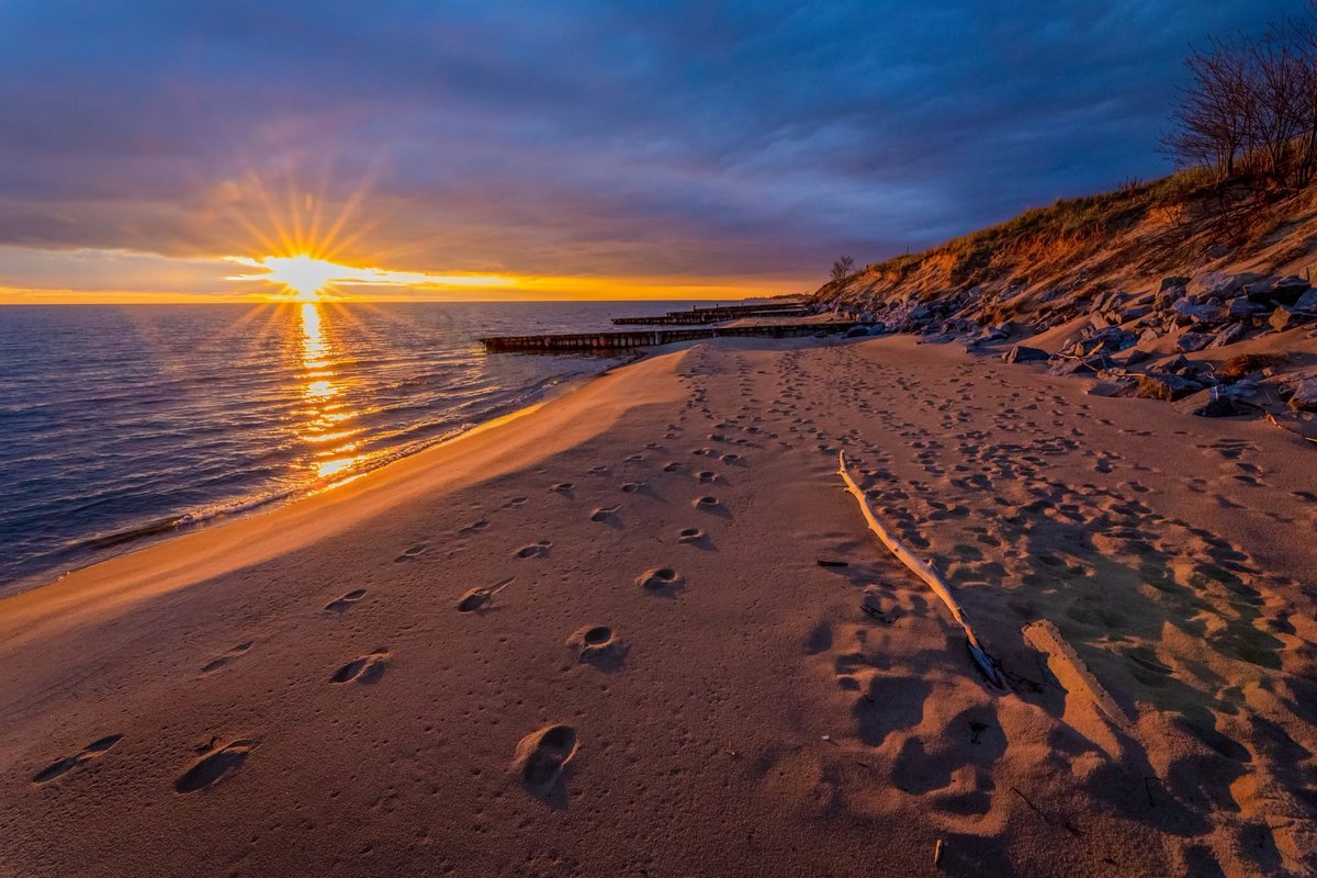 Sun sets on Lake Michigan with footprints lining the sandy shoreline.