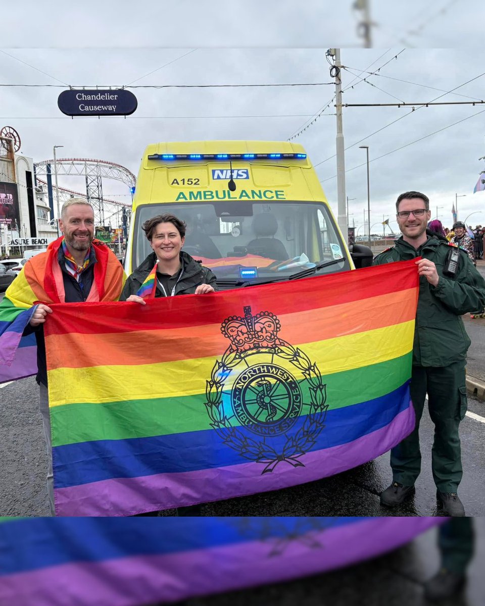 Three NWAS colleagues stand in front of an ambulance holding a rainbow NWAS Pride flag at Blackpool Pride.