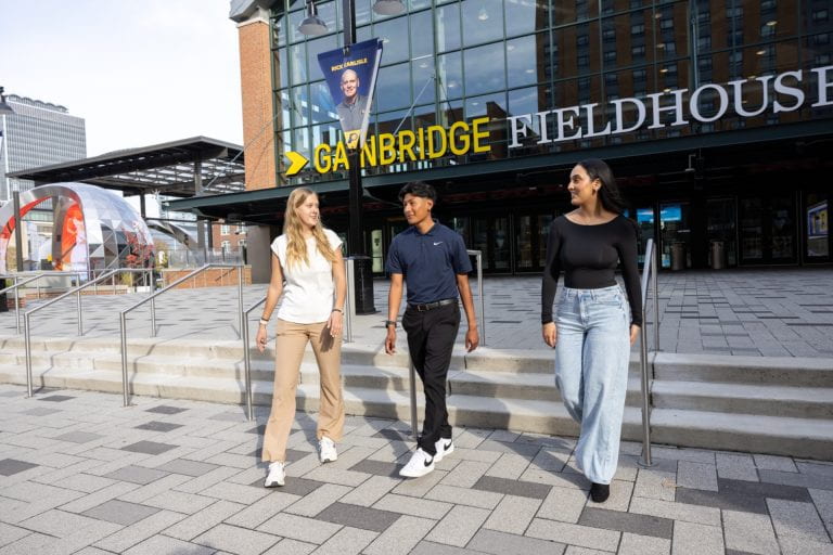 Two people walk with a third person outside the Gainbridge Fieldhouse. They appear to be engaged in conversation. The background features the entrance to the building with a banner displaying Larry Bird's image.