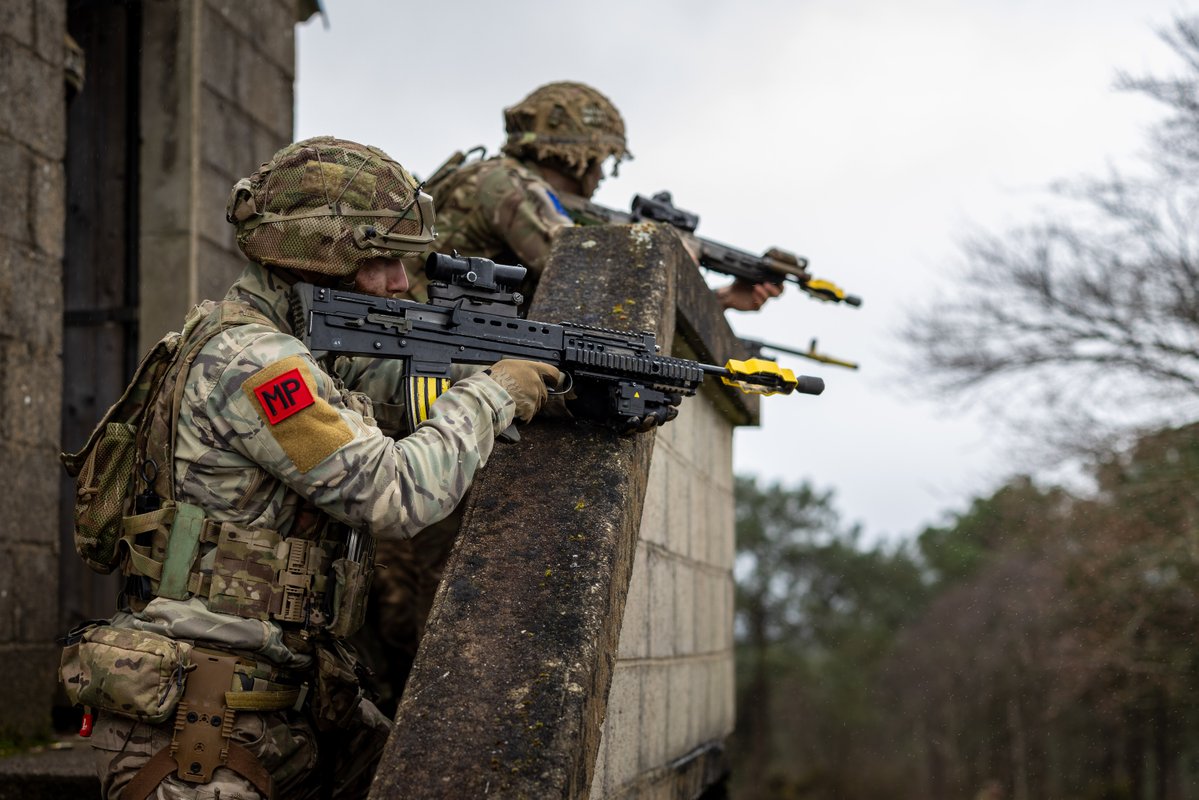Two armed soldiers in camouflage uniforms and helmets are positioned on a rooftop ledge, aiming rifles into the distance. One soldier has an "MP" patch on their sleeve. The background includes a stone building and trees under a cloudy sky.
