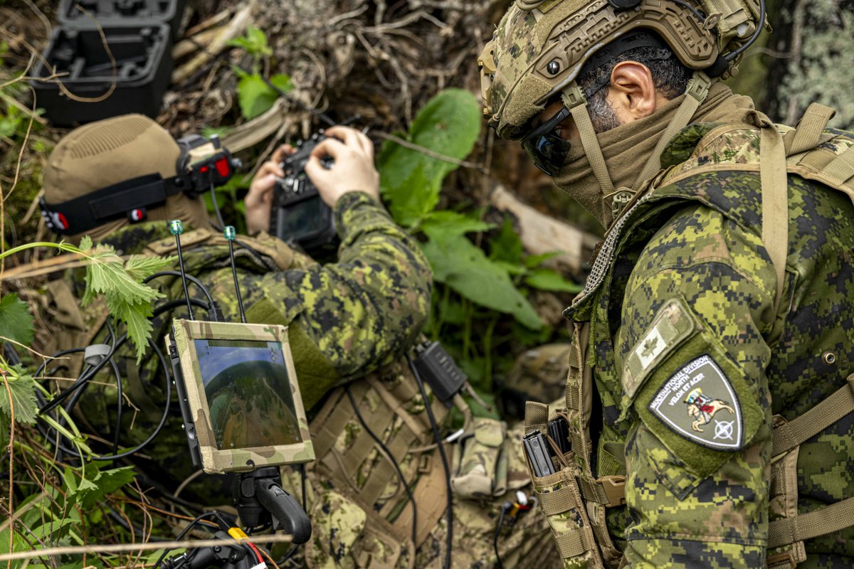 Deux soldats en tenue de camouflage utilisent de l’équipement électronique en milieu forestier dense, en se concentrant sur un appareil doté d’un écran et d’une antenne.